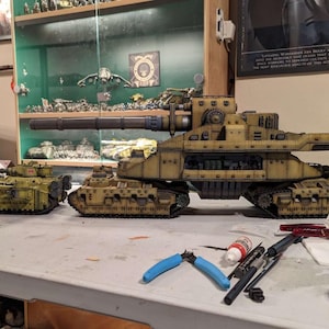 May include: A collection of model tanks in various sizes, painted in shades of tan and olive green. The largest tank has a long cannon and detailed mechanical features. Tools and supplies are scattered on the table, suggesting a hobbyist's workspace.