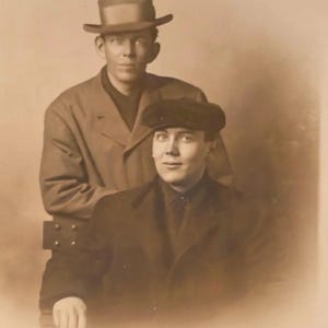 Puede incluir: Retrato en tonos sepia de dos hombres. Uno de ellos está de pie detrás del otro, con un abrigo largo y un sombrero fedora. El hombre de delante está sentado, con un abrigo oscuro y una gorra plana. La fotografía tiene una estética vintage.