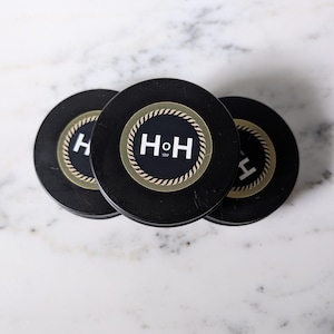 May include: Three black coasters with a white and gold rope design around the edge. The center of each coaster features the white letters "H" and "H" with "SHOP" below. The coasters are arranged on a white and gray marble surface.