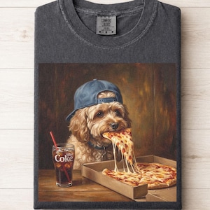 May include: A charcoal gray t-shirt featuring a graphic of a fluffy dog wearing a blue baseball cap, eating a slice of pizza from a cardboard box, and a glass of Diet Coke with a red straw. The shirt has a Comfort Colors brand neck label.