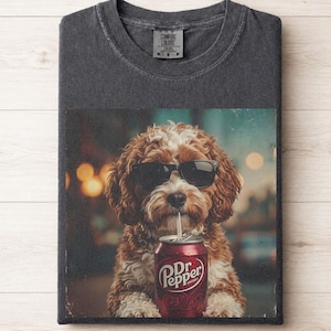 May include: A charcoal gray t-shirt featuring a graphic print of a brown and white curly-haired dog wearing black sunglasses and drinking from a red Dr Pepper soda can. The shirt has a Comfort Colors brand label at the inner neckline.