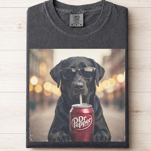 May include: A charcoal gray crew neck t-shirt featuring a graphic print of a black Labrador dog wearing black sunglasses and drinking from a red Dr. Pepper soda can with a straw. The shirt has a Comfort Colors brand label at the inner neckline.