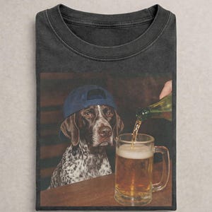 May include: A charcoal gray t-shirt featuring a graphic print of a brown and white spotted dog wearing a blue baseball cap. The dog is positioned next to a glass beer mug being filled with a golden liquid from a green glass bottle.