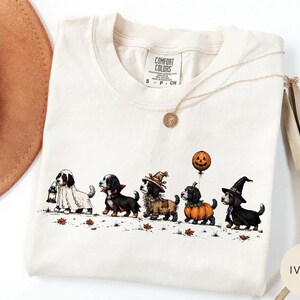 May include: An ivory t-shirt with a Comfort Colors label in size S. The graphic features five dogs in Halloween costumes: a ghost with a lantern, a vampire, a scarecrow, a dog in a pumpkin suit with a balloon, and a witch. Scattered autumn leaves.