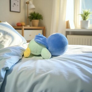 Puede incluir: Un peluche en tonos de azul, verde y amarillo descansa sobre una cama cubierta con una s&aacute;bana azul claro. El peluche tiene una forma redondeada con una cabeza azul y un cuerpo verde claro, creando una est&eacute;tica suave y acogedora.