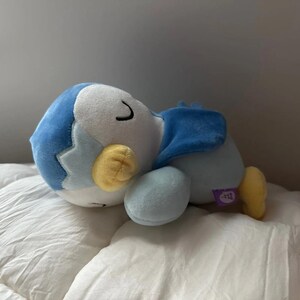 Puede incluir: Un peluche de un ping&uuml;ino durmiendo, con cuerpo azul y blanco, pies y pico amarillos. El ping&uuml;ino est&aacute; acostado sobre una superficie blanca. El juguete tiene una peque&ntilde;a etiqueta morada.