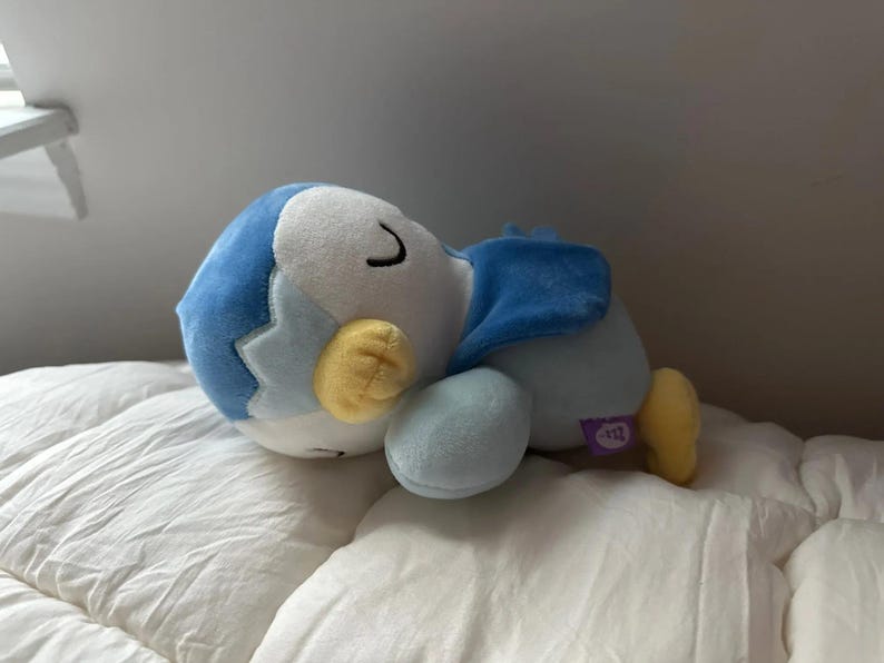 Puede incluir: Un peluche de un ping&uuml;ino durmiendo, con cuerpo azul y blanco, pies y pico amarillos. El ping&uuml;ino est&aacute; acostado sobre una superficie blanca. El juguete tiene una peque&ntilde;a etiqueta morada.