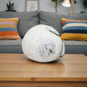 May include: A plush, round seal pillow in a sleeping position. The pillow is primarily white with gray spots and a detailed face. The seal is resting on a wooden table, with a gray couch and colorful pillows in the background.