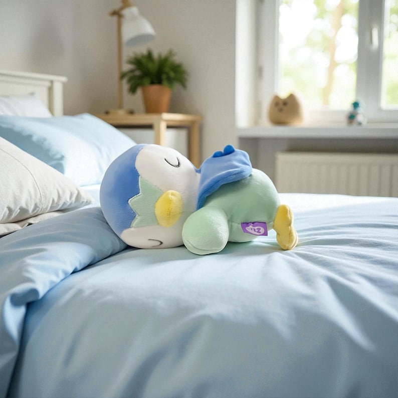 Puede incluir: Un peluche con forma de ping&uuml;ino, con colores azul, verde, amarillo y blanco, descansa sobre una cama azul claro. El ping&uuml;ino est&aacute; tumbado, con la cabeza apoyada en el brazo. El juguete tiene un aspecto suave y tierno.
