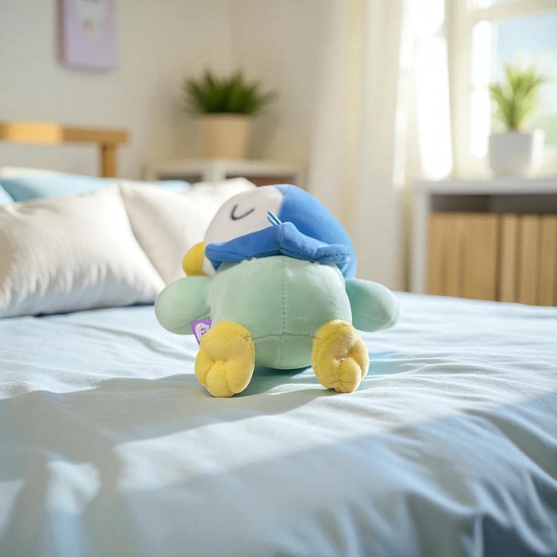 Puede incluir: Un peluche de ping&uuml;ino en tonos azules, verdes y amarillos est&aacute; sobre una colcha azul claro. El ping&uuml;ino tiene un gorro azul, pies amarillos y un cuerpo verde claro. El juguete es suave y tierno.