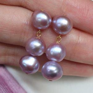 Op de afbeelding: Een paar lavendelkleurige parel oorbellen. Elke oorbel heeft drie glanzende, ronde parels die aan een goudkleurige haak hangen. De parels hebben een zachte, iriserende glans, die licht reflecteert met hints van roze en wit.