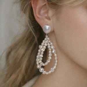 May include: Elegant white pearl earrings. Each earring features a large, round pearl stud, with a cascading teardrop design of smaller, irregularly shaped pearls. The pearls have a lustrous, creamy white color, creating a sophisticated look.