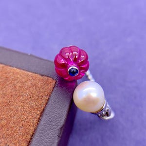 May include: A silver ring with a carved fuchsia gemstone, a small blue stone, and a white pearl. The ring is set against a purple background.