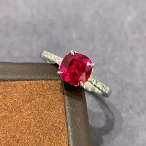 May include: A silver ring with a large, square-cut, vibrant red gemstone. The band is adorned with small, clear accent stones. The ring is displayed on a brown surface against a gray background.