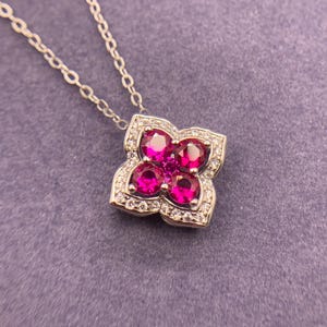 May include: A silver necklace with a flower-shaped pendant. The pendant features four large, round, bright pink gemstones surrounded by smaller clear stones. A smaller pink gemstone is in the center of the flower. The chain is delicate and silver.