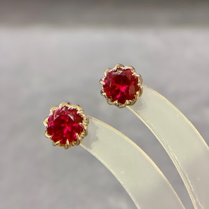 May include: A pair of stud earrings featuring a vibrant red gemstone in a floral setting. The earrings have a gold-colored metal border, and the gemstones are faceted to catch the light. The earrings are displayed on a clear, curved stand.