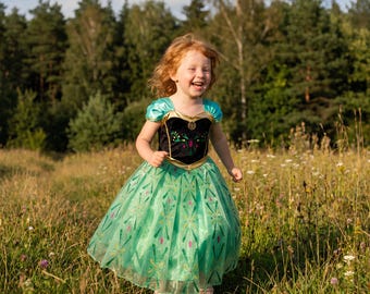 Op de afbeelding: Een kind draagt een jurk met een zwart fluwelen lijfje, gouden randen en korte turquoise mouwen. De rok is gemaakt van lichtgroene tule met een glinsterend patroon van roze en gouden ruitvormen en lijnen door de hele stof.