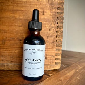Peut inclure: Une bouteille en verre brun d'élixir de sureau avec un compte-gouttes noir. L'étiquette indique "Ambrose Apothecary" et "élixir de sureau". La bouteille contient 60 ml de liquide. La bouteille est posée sur un fond en bois.