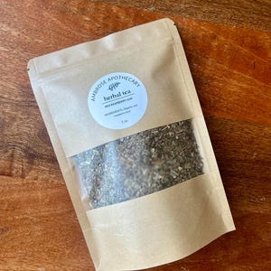 May include: A kraft paper pouch of herbal tea with a clear window, displaying the tea leaves. The label reads "Ambrose Apothecary, herbal tea, Red Raspberry Leaf, Ingredients: Organic red raspberry leaf, 1 oz."