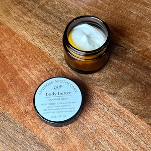 Op de afbeelding: Een klein, open amberkleurig glazen potje gevuld met witte body butter, naast een potdeksel. Het deksel is zwart met een lichtblauw etiket met de tekst "AMBROSE APOTHECARY body butter SIGNATURE SCENT" en informatie over de ingrediënten. De pot bevat 60 ml.