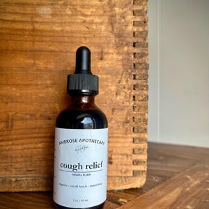 Op de afbeelding: Een donker amberkleurige glazen fles met een zwarte druppelaar, met het label "AMBROSE APOTHECARY cough relief HERBAL ELIXIR." Het label vermeldt ook "organic - small batch - nourishing" en "2 oz. / 60 ml." De fles staat tegen een houten achtergrond.