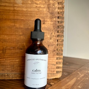 May include: A brown glass bottle of "calm" herbal tincture from Ambrose Apothecary. The label is white with black text and a botanical illustration. The bottle has a black dropper top and contains 60 ml of liquid.