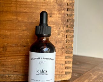 Puede incluir: Una botella de vidrio marrón de tintura herbal "calm" de Ambrose Apothecary. La etiqueta es blanca con texto negro y una pequeña ilustración botánica. La botella tiene un tapón cuentagotas negro y contiene 60 ml de líquido.