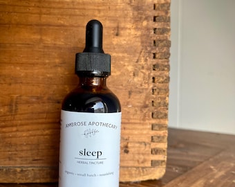 Pode incluir: Uma garrafa de vidro castanho com um conta-gotas preto, rotulada como "Ambrose Apothecary Sleep Herbal Tincture". O rótulo também indica "organic - small batch - nourishing" e contém 60 ml. A garrafa está sobre uma superfície de madeira.