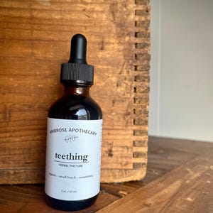 May include: A dark amber glass bottle with a black dropper, labelled "teething" by Ambrose Apothecary. The label also states "herbal tincture" and "60 ml". The bottle is set against a wooden background.