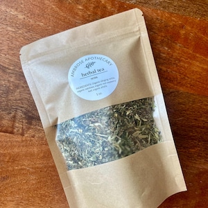 Può includere: Una busta di carta kraft di tè alle erbe "NORA" di Ambrose Apothecary. La finestra trasparente rivela erbe essiccate. L'etichetta elenca gli ingredienti: ortica biologica, avena, foglie di lampone rosso e erba medica. La busta contiene 28 grammi.