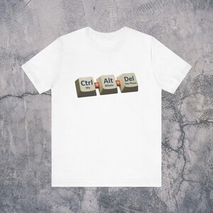 Pode incluir: Camiseta branca com um gráfico de três teclas de computador: Ctrl, Alt e Del. O texto nas teclas diz "Ctrl My", "Alt Whole" e "Del Day Please". A camisa é feita de um material macio.