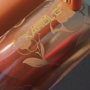 May include: A metallic copper-colored tumbler featuring a glittery finish and an etched floral design. The text KAROL G is engraved in a stylized font across the surface, integrated into the floral pattern. The object has a smooth, reflective texture.