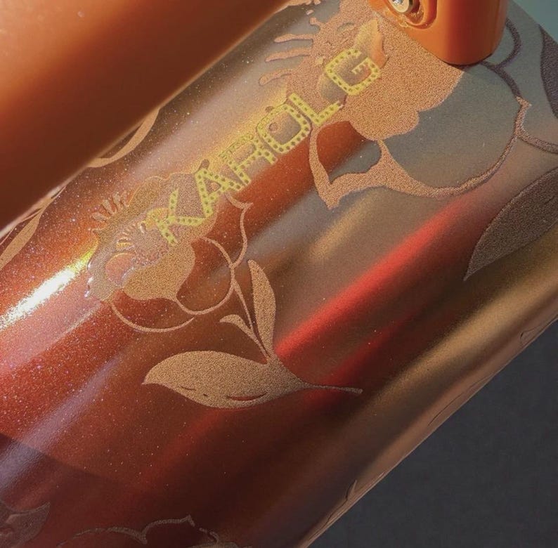 May include: A metallic copper-colored tumbler featuring a glittery finish and an etched floral design. The text KAROL G is engraved in a stylized font across the surface, integrated into the floral pattern. The object has a smooth, reflective texture.