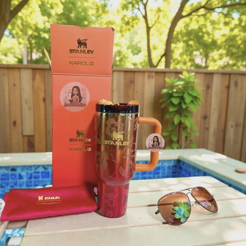 May include: A metallic red Stanley Karol G tumbler with a floral pattern and orange handle, standing next to its matching orange box. A red Stanley branded pouch and brown aviator sunglasses with a small green floral embellishment are also present.