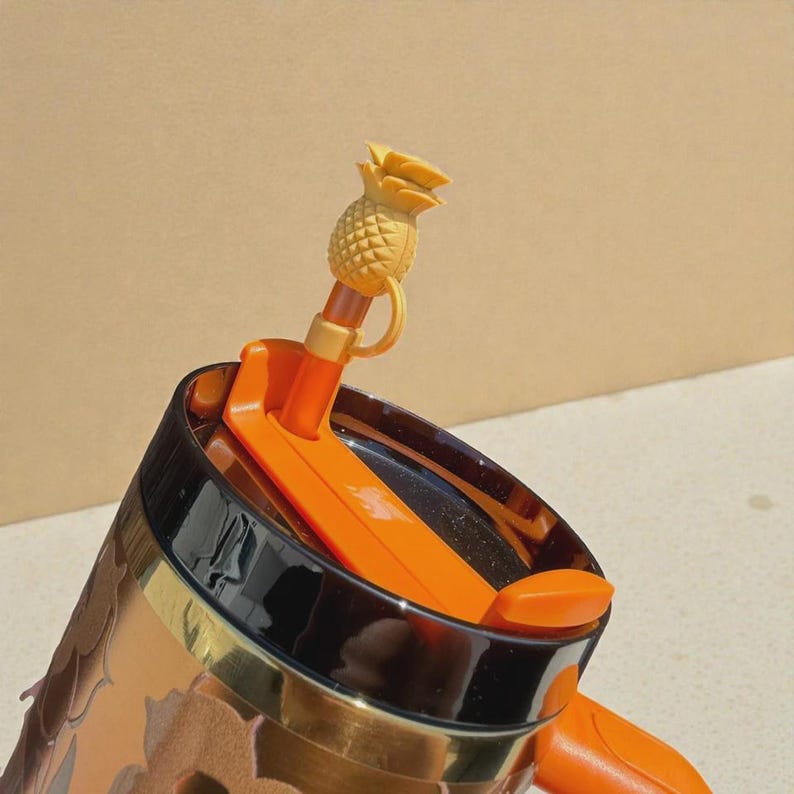 May include: A dark-colored tumbler featuring a black rim and an orange lid. A straw is inserted into the lid, topped with a small, textured, yellow-orange pineapple-shaped silicone cover attached by a flexible loop to the straw.