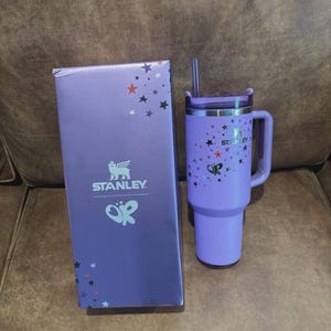May include: A light purple Stanley tumbler with a matching straw and handle, featuring a pattern of small stars and a butterfly graphic. Beside it stands a rectangular purple box displaying the Stanley logo, a butterfly icon, and star accents.