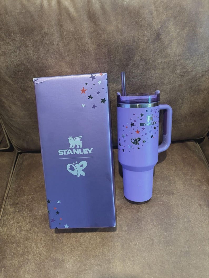 May include: A light purple Stanley tumbler with a matching straw and handle, featuring a pattern of small stars and a butterfly graphic. Beside it stands a rectangular purple box displaying the Stanley logo, a butterfly icon, and star accents.