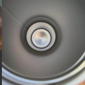 May include: A top-down view into the interior of a cylindrical container with a smooth, matte grey finish. The base features a circular, metallic silver center disc with a small, embossed logo detail, surrounded by concentric rings.