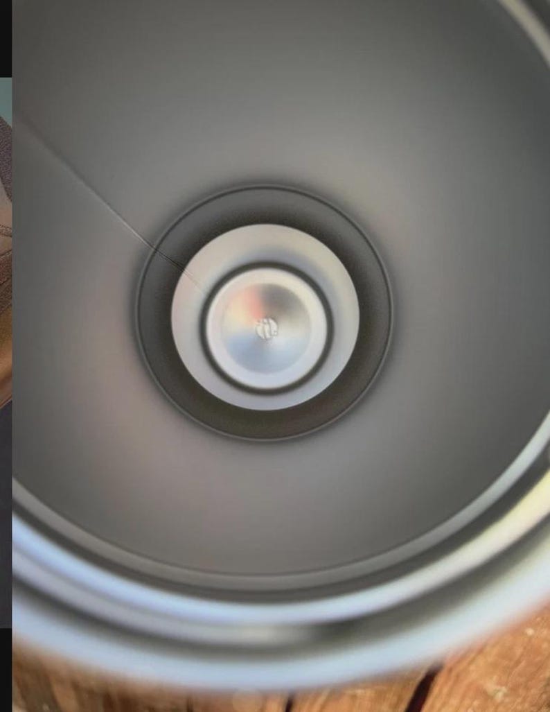 May include: A top-down view into the interior of a cylindrical container with a smooth, matte grey finish. The base features a circular, metallic silver center disc with a small, embossed logo detail, surrounded by concentric rings.