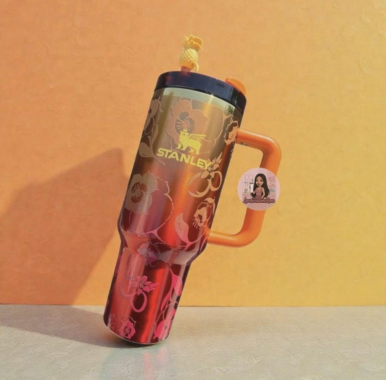 May include: A Stanley brand tumbler with a dark brown and orange gradient finish featuring a floral pattern. The cup has a sturdy orange handle, a dark lid, and a small yellow pineapple-shaped straw topper. A circular sticker with a cartoon character is attached to the handle.