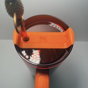 May include: A dark translucent brown tumbler with a bright orange lid and handle. The lid features a central orange tab with a small embossed winged lion logo and a circular opening holding a clear straw topped with a gold-colored pineapple charm.