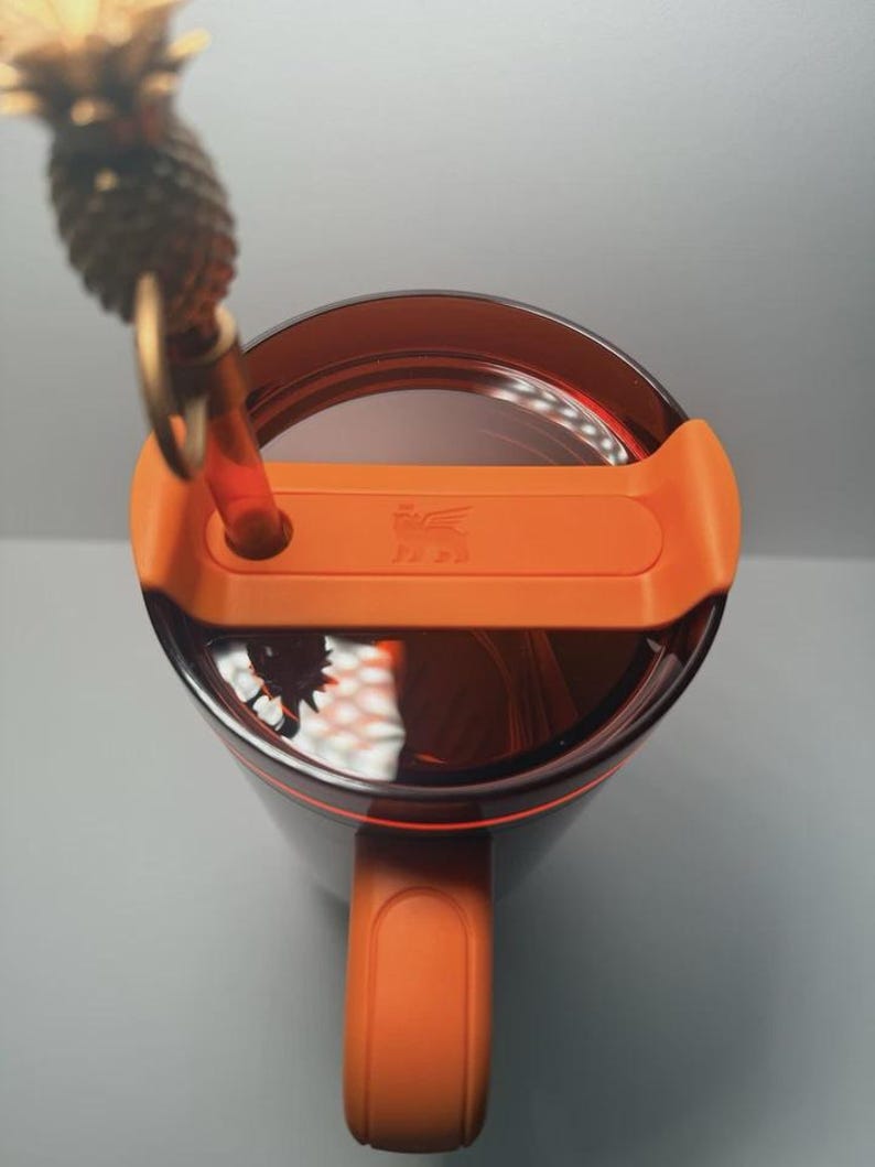 May include: A dark translucent brown tumbler with a bright orange lid and handle. The lid features a central orange tab with a small embossed winged lion logo and a circular opening holding a clear straw topped with a gold-colored pineapple charm.