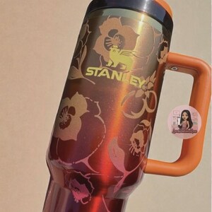 May include: A metallic red Stanley tumbler featuring a dark brown lid, an orange handle, and a floral pattern in shades of gold and pink. The Stanley logo is printed in gold on the upper body, and a small circular sticker is attached to the handle.