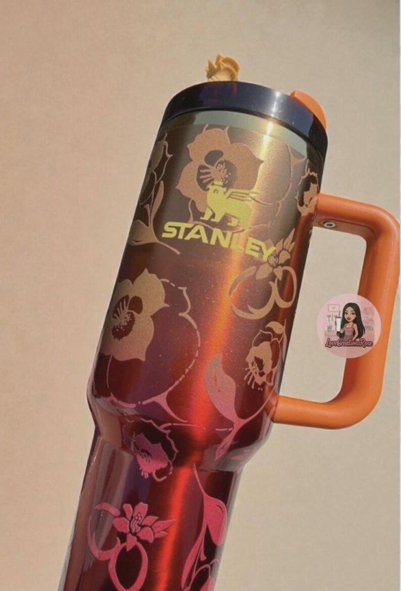 May include: A metallic red Stanley tumbler featuring a dark brown lid, an orange handle, and a floral pattern in shades of gold and pink. The Stanley logo is printed in gold on the upper body, and a small circular sticker is attached to the handle.