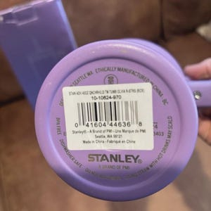 May include: A lavender-colored Stanley brand tumbler with a circular base. The bottom features a white product label with a barcode, the text STAN ADV 40OZ QNCHRH2.0 TM TUMB OLVIA R-STRS (BCX), and the model number 10-10824-970 printed on it.