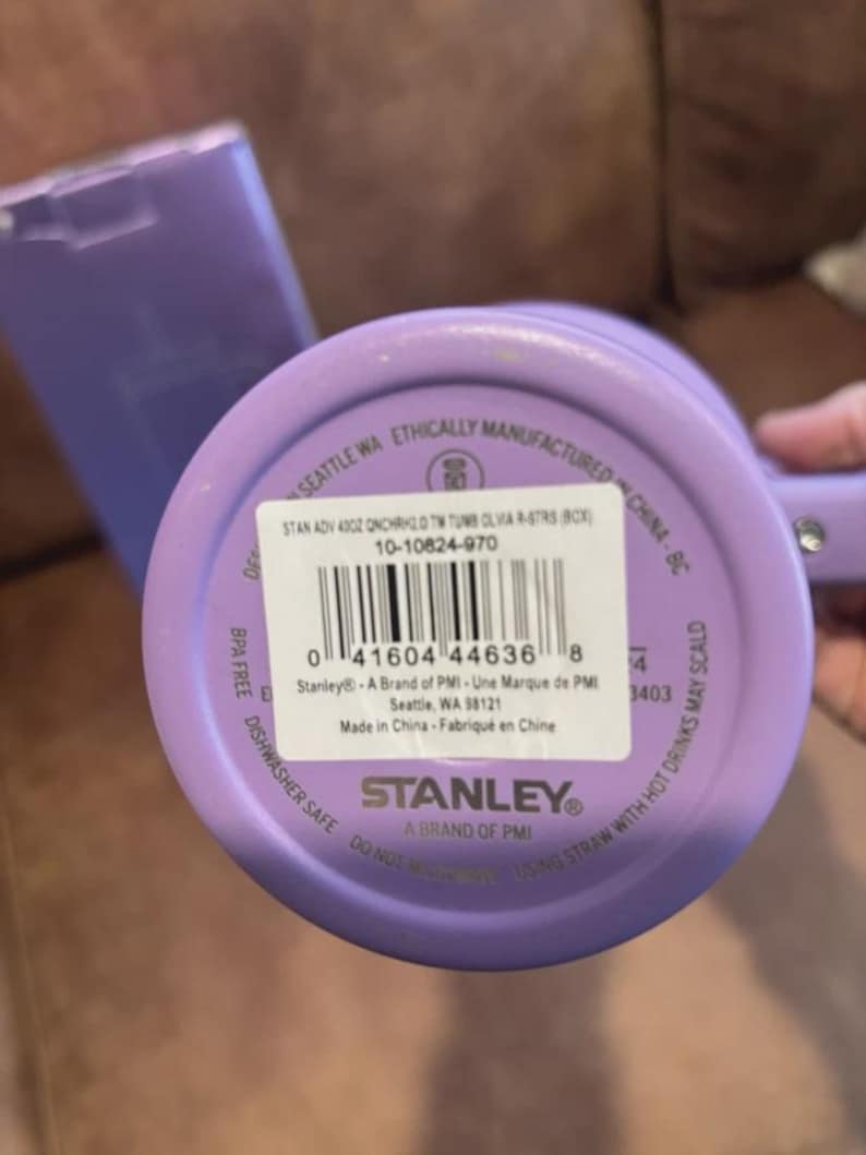 May include: A lavender-colored Stanley brand tumbler with a circular base. The bottom features a white product label with a barcode, the text STAN ADV 40OZ QNCHRH2.0 TM TUMB OLVIA R-STRS (BCX), and the model number 10-10824-970 printed on it.