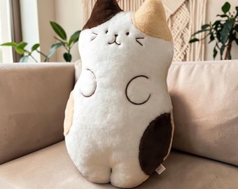 May include: A soft cat-shaped cushion in white, brown, and tan. The cushion features a smiling face with embroidered details and a rounded body. The cat has brown spots and a tan ear. The cushion is on a beige sofa.