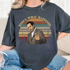 May include: Dark gray t-shirt featuring a retro design with the text "HERE'S WHAT HAPPENED." The design includes an image of a man in a suit with his hand raised, set against a sunset-colored background.