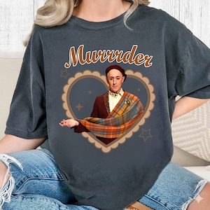 May include: Dark grey t-shirt with the word "Murrrder" in brown script. The shirt features a heart-shaped graphic with a portrait of a man in a beret and plaid shawl. The graphic is surrounded by stars.