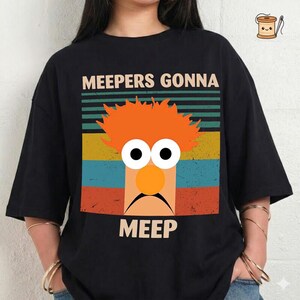 May include: Black t-shirt featuring a colorful graphic of a cartoon character with orange hair, large eyes, and a round orange nose. The text "MEEPERS GONNA MEEP" is printed above and below the character in a bold font.